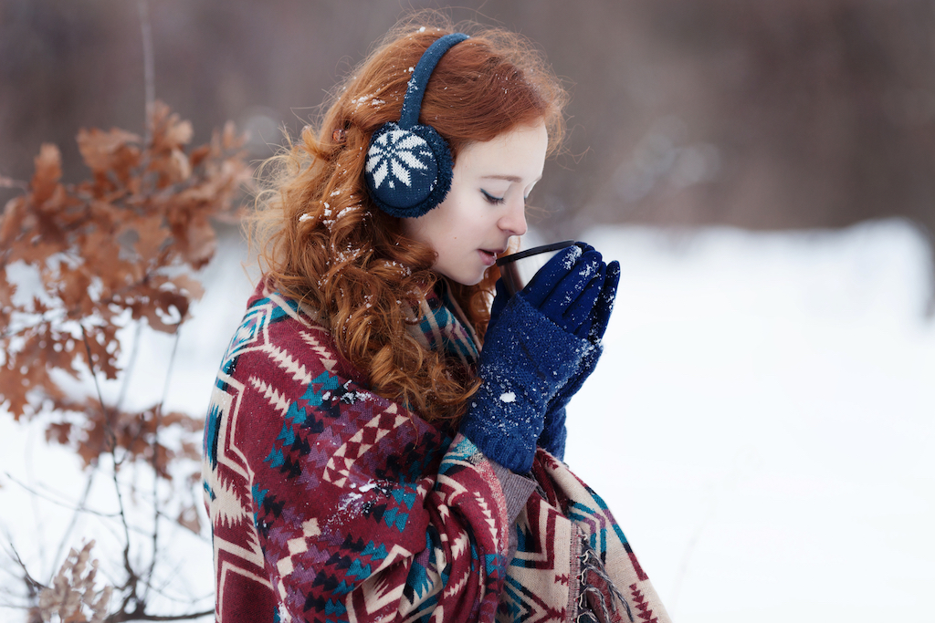 Attractive young red-haired woman drinking a hot drink from a mug in the winter park. She is wearing a blue headset and gloves and a scarf with ethnic patterns.